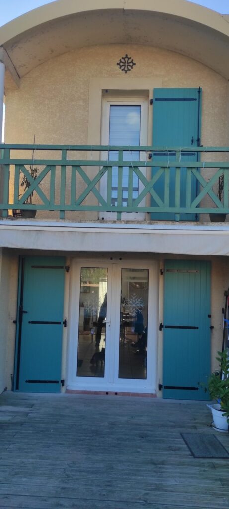 Volets turquoise et balcon pour maison avec croix occitane Maison avec volets turquoise, balcon sculpté et terrasse bois, sous une croix occitane.
