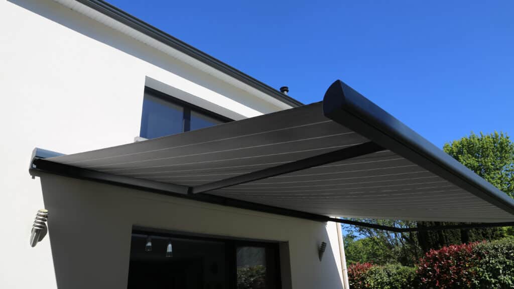 Store banne motorisé gris : protection solaire terrasse Store banne motorisé gris anthracite sur façade de maison moderne sous un ciel bleu.